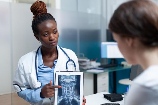 African american radiologist doctor discussing bones radiography with woman patient explaining medical expertise during clinical appointment in hospital office. Radiology on tablet computer screen