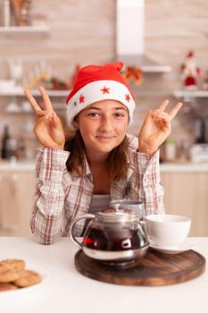 Portrait of child wearing santa hat looking into camera standing at table in xmas decorated kitchen. Girl enjoying winter season celebrating christmas holiday. Baked traditional cookies on table