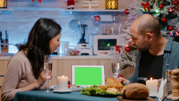 Couple looking at horizontal green screen on tablet while eating meal at festive dinner on christmas eve. Cheerful people watching chroma key with isolated mockup template enjoying festivity