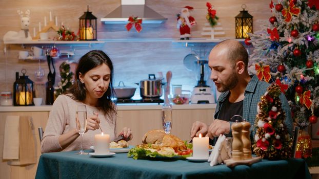 Cheerful couple celebrating christmas eve with festive dinner. People enjoying seasonal meal with glasses of champagne at home for winter holiday celebration. Man and woman eating food