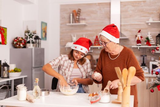 Grandchild mixing ingredients in bowl preparing traditional homemade dough with grandmother celebrating christmas season together in xmas decorated kitchen. Kid enjoying winter holiday baking cookies