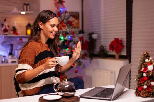 Cheerful person holding cup of tea while on video call for online conference on laptop computer in festive kitchen at home. Caucasian young woman enjoy talking to family on christmas
