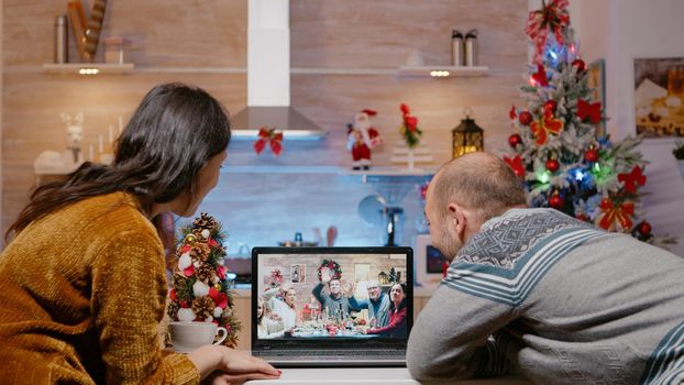 Couple using video call talking to family on christmas eve. Festive man and woman celebrating holiday festivity with relatives on online remote conference on laptop. Christmas dinner on internet