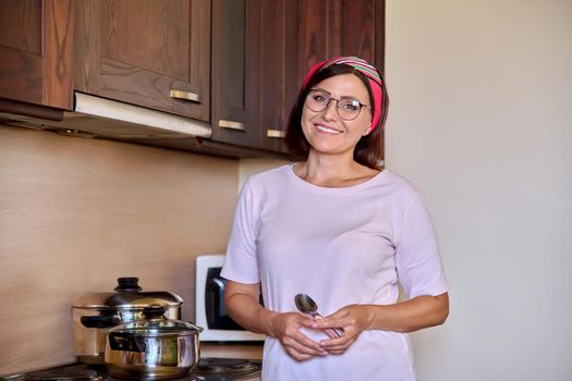 Portrait of middle aged smiling woman preparing food in kitchen. Mature female wearing headband glasses looking at the camera, furniture hob with pans background. Cooking, homemade food, lifestyle