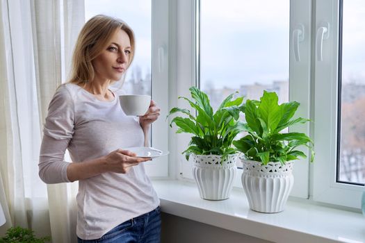 Middle aged beautiful woman with a cup of coffee at home near the window in the winter autumn season, copy space. Beauty, lifestyle, comfort, tranquility, mature age concept