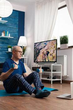 Retired senior man sitting on yoga mat stretching abdominals muscles practicing fitness workout using dumbbells. Pensioner working at body warming up during wellness routine watching pilates video