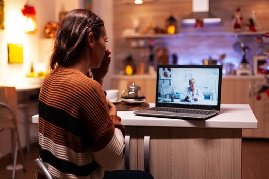 Sick woman talking to doctor on online video conference while sitting at home in festive kitchen. Unwell caucasian patient using telemedicine for prescription treatment and advice