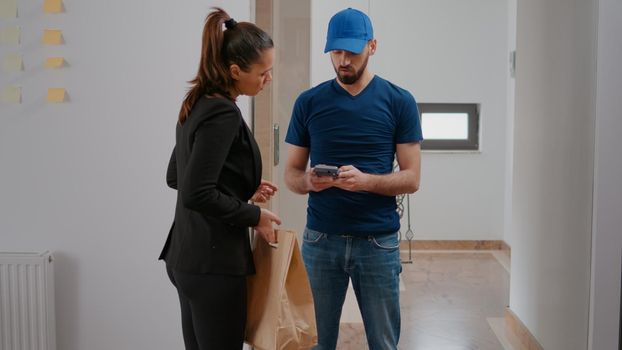 Businesswomn working in startup company office receiving takeaway food meal box paying order with smartwatch using POS service. Delivery guy worker delivering lunch purchase during lunchtime