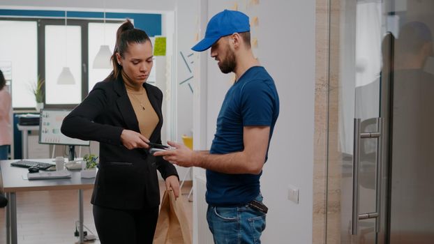 Businesswoman receiving meal order package paying food transaction with contactless credit card using POS. Delivery man delivering takeaway lunch box order in business company office