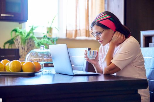 Middle-aged woman sitting at home in kitchen with laptop and cup of coffee. Lifestyle, leisure, technology, work, relaxation, mature people concept