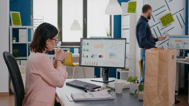 Executive manager eating tasty sandwich while analyzing marketing strategy at desk in workplace. Takeout order food delivery in corporate place, lunch meal break package delivered at startup office.