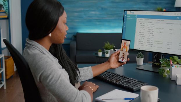 Student sitting at desk in living room having online videocall teleconference meeting using phone webcam. Woman discussing with remote team explaining marketing college course using elearning platform