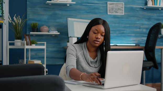 African american student working remote from home at marketing strategy writing financial graphs on notebook. Black woman sitting on bean bag in living room learning company statistics