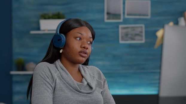 African american student putting headphones enjoying music while working remote from home searching online course using university platform. Black woman sitting at desk in living room drinking coffee