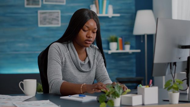 Black student sitting at desk writing school homework on notebook during online courses education. Young woman browsing communication project on computer in living room typing webinar notes