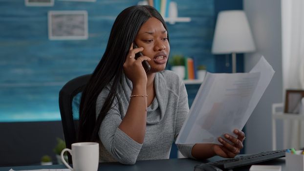 Upset black student discussing with collegue at phone analyzing paperwork with financial course sitting at desk working remote from home. Woman doing university homework using elearning platform