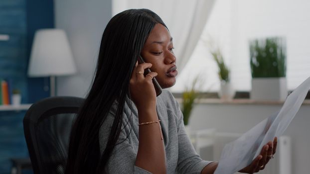 Upset black student sitting at desk explaining financial homework discussing with collegue using smartphone. Black woman working remote from home at online webinar using academic elearning platform