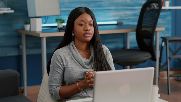 African american student with dark skin waving school team working from home at marketing course during online videocall conference meeting. Computer user sitting on bean bag in living room