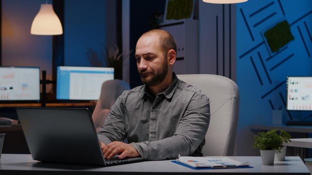 Overworked businessman typing financial statistics on laptop sitting at desk table in startup company office late at night. Workaholic entrepreneur planning economic project before company deadline