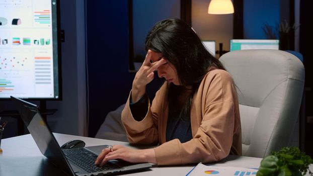 Overworked tired businesswoman rubbing face working at management statistics on laptop computer late at night in startup office. Exhausted entrepreneur checking economic graphics company deadline