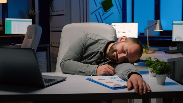Exhausted overworked businessman sleeping on desk table in startup business office after planning management statistics late at night. Workaholic entrepreneur working at economic deadline project