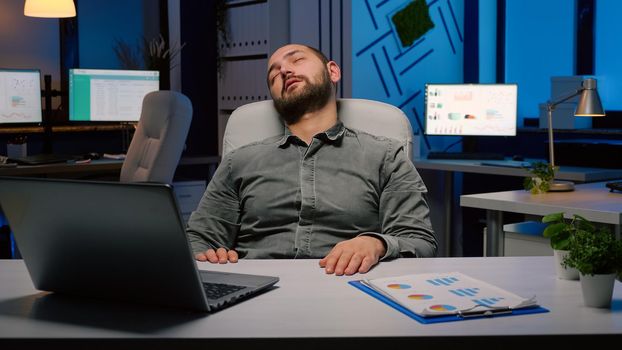 Workaholic businessman slepping on chair in business company office late at night after checking financial statistics on laptop computer. Exhausted manager working overworked at economic project