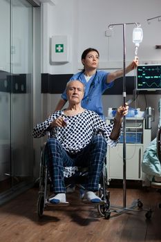 Medical practitioner preparing senior sick hospitalized man in wheelchair, checking iv drip. Nurse with stethoscope preparing patient for examinantion. Oximeter attached to finger of man.