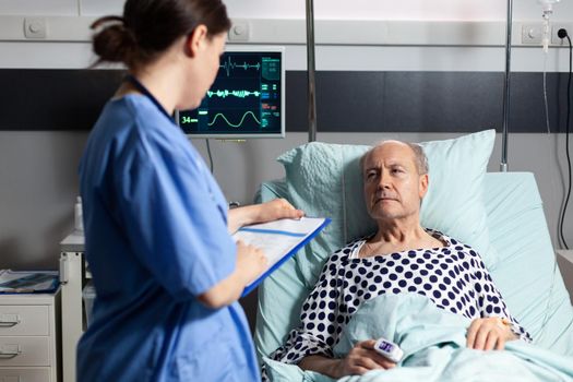 Medical assistant checking treatment of senior man reading notes on clipboard. Sick elderly laying in bed discussing with nurse about diagnosis expertise with iv bag attached.