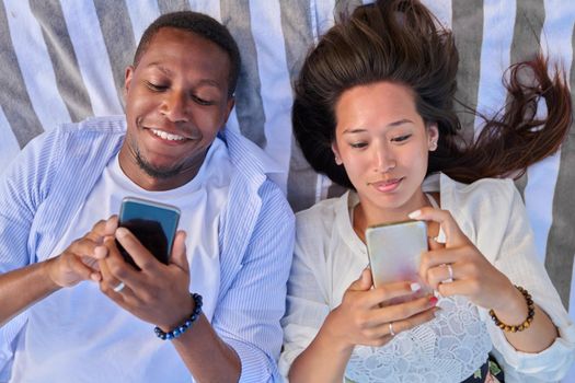 Top view smiling young couple looking at smartphone screens. Multiracial couple of asian woman and african man outdoor vacationing, lying with phones in hand. Lifestyle, leisure, technology, people