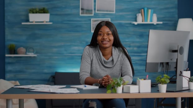 Portrait of smiling student looking into camera while sitting at desk table in living room during home school. Black woman studying highschool news browsing academic information on computer