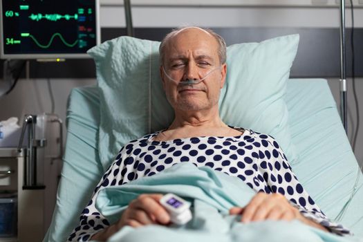 Portrait of sick senior man patient resting in hospital bed, breathing with help from oxygen mask because of lungs infection, having oximeter attached on finger.