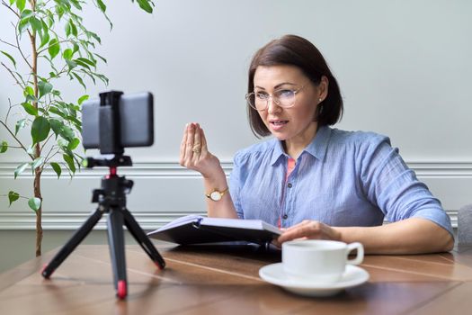 Middle-aged woman teacher, business woman working online using smartphone. Business confident female with notepad on table talking looking in webcam phone on tripod, at home. Technology education work