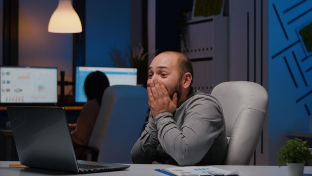 Happy cheerful businessman smiling while looking at laptop computer display receiving online good job promotion. Joyful happy executive manager working in company office late at night