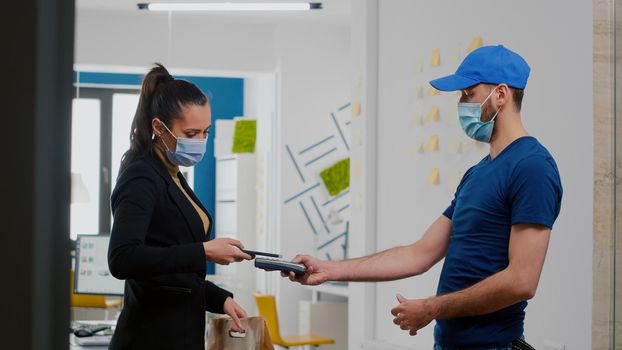 Businesswoman with face mask receiving takeaway food lunch order paying with smartphone using POS contactless service. Delivery man with gloves against covid19 bringing fresh lunch at office workplace