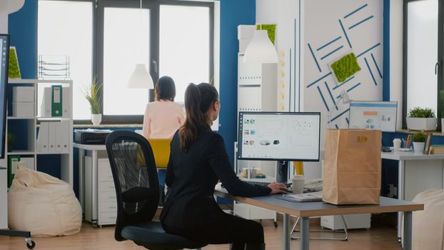 Executive manager holding takeaway food bag order during takeout lunchtime in startup business corporate office. Businesswoman sitting at desk table during lunch break working at financial graphs