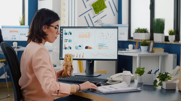 Businesswoman having delivery lunch food order during takeaway lunchtime working in business company office. Executive manager woman eating takeaway sandwich meal typing management statistics