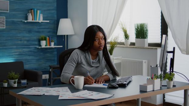 Black student drinking coffee while searching business information on internet using computer working at communication project. Focused woman sitting at desk in living room analyzing homework graphic