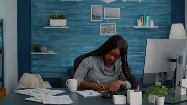 Black student writing school ideas on stickey notes working at homework sitting at desk table in living room. Young woman studying business at univeristy using elearning platform during online course