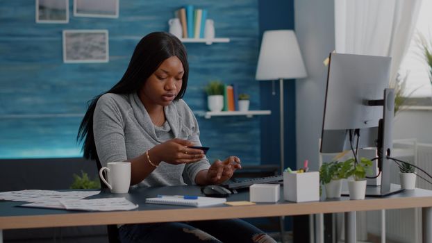 African american student holding cred card in hands doing digital transcation browsing online store working remote from home. Teenager searching banking service on computer in living room