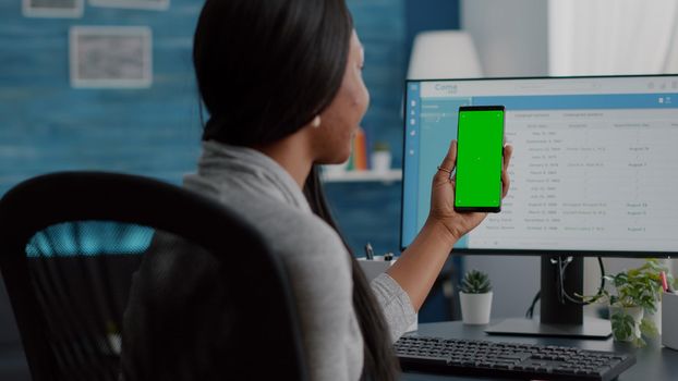 African woman analyzing social media video using mock up green screen chroma key phone with isolated display working remote from home sitting at desk in living room. Student browsing social network