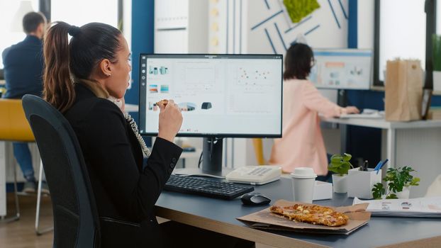 Multitasking businesswoman discussing marketing strategy with remote worker using landline while eating pizza slice during lunchtime. Lunch meal takeaway order package delivered at startup office