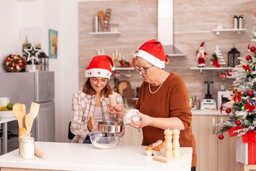 Family celebrating christmas holiday preparing homemade dough dessert in xmas decorated culinary kitchen at home. Grandma putting flour in strainer while child shacking enjoying winter season