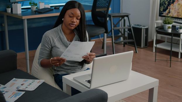 African american student analyzing financial paperwork graph typing infographics presentation working remote from home sending email to company. Black woman sitting on bean bag in living room