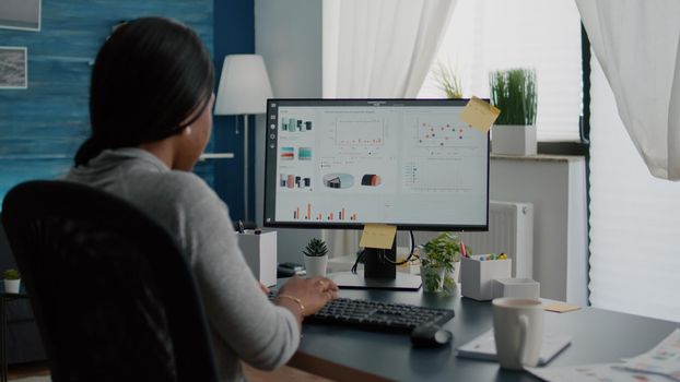 Closeup of black student browsing financial infographics analyzing statistics on laptop computer working at high school webinar. Woman sitting at desk table in living room typing commerce chart graph