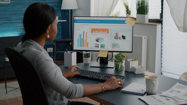 High school black student typing financial information analyzing digital statistics using university online webinar. Smart woman sitting at desk in living room browsing commerce chart on computer