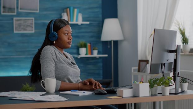 Student with black skin having headphone puts listening online univeristy course using elearning platform sitting at desk in living room. African american woman working remote from home