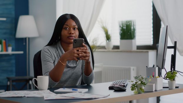 Black student holding phone in hands chatting with people browsing communication information sitting at desk in living room. Teenager looking on social media sharing lifestyle advice to group friends