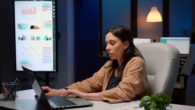 Exhausted businesswoman analyzing marketing statistics on laptop computer working in startup business company office. Tired workaholic manager remains alone in financial room after his collegue left