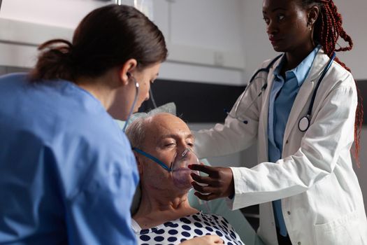 Side view of senior patient breathing assisted by respiratory tube in hospital intesive care. African doctor and nurse helping old man to breath sing face mask.