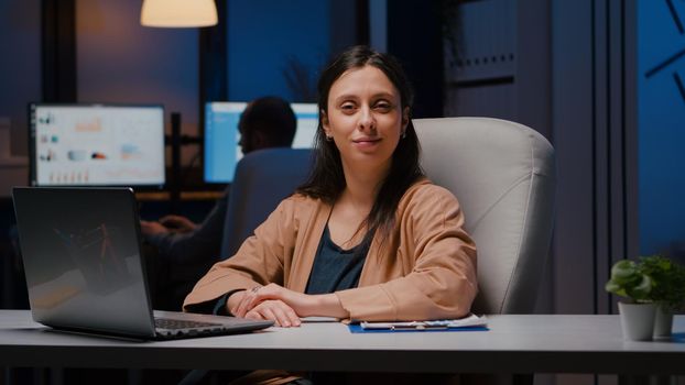 Portrait of smiling businesswoman looking into camera while sitting at desk in startup business office late at night. Manager woman planning financial statistics working at marketing statistics